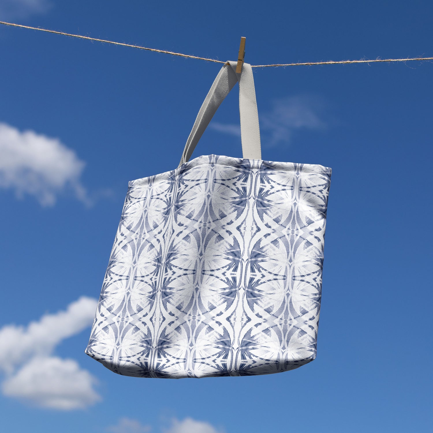 Large tote bag featuring a blue and white abstract pattern, hanging from a clothesline with a bright blue sky background.