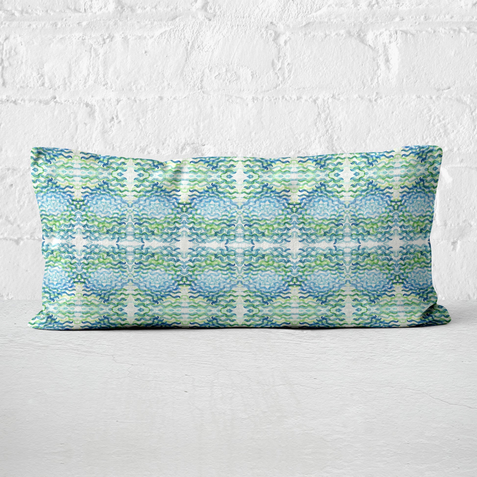 12x24 lumbar pillow featuring an abstract hand-painted pattern in green and blue leaning against a white brick wall.