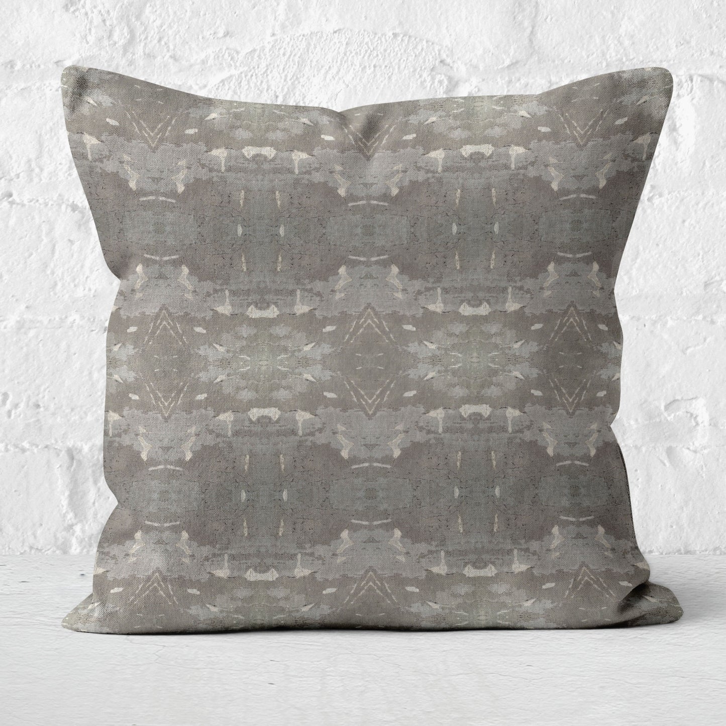 Square throw pillow featuring an abstract gray and khaki pattern, sitting in from of a white brick wall