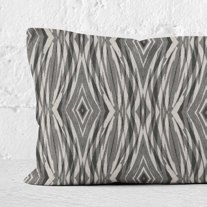 Close up of a rectangular lumbar pillow featuring linocut pattern in grey and neutral tones leaning against a white brick wall.