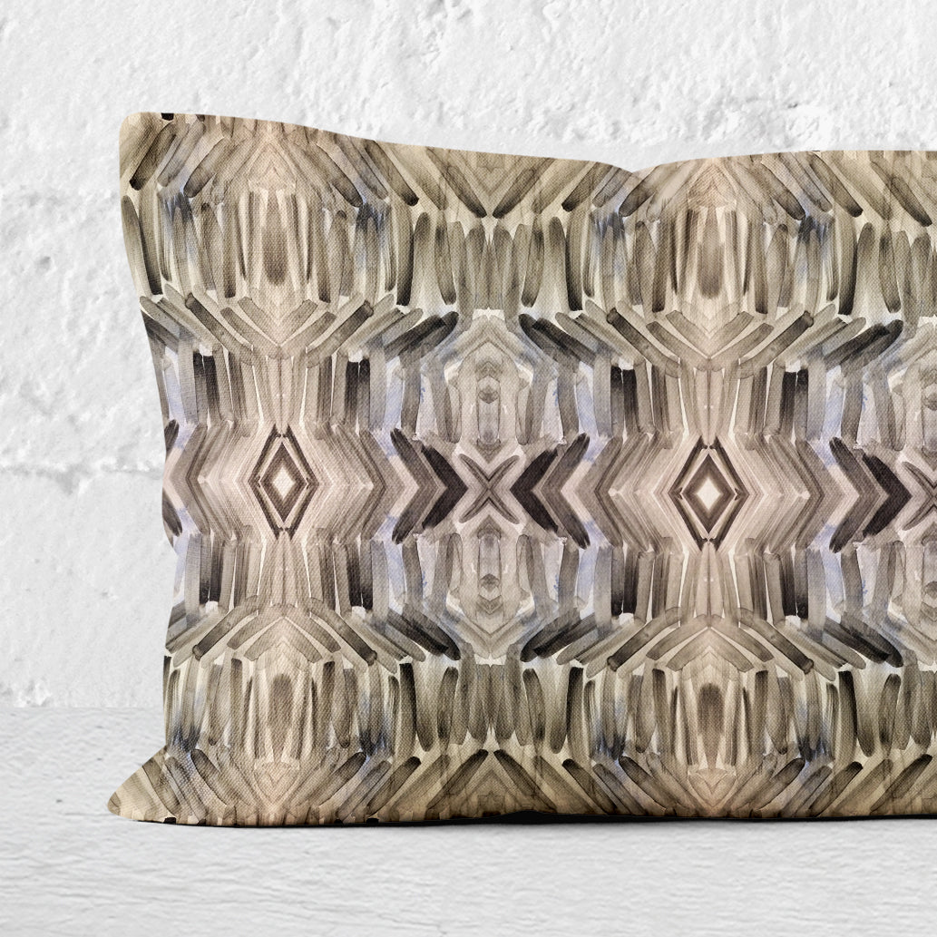 Detail of a 12x24 rectangular pillow featuring a brown abstract hand-painted pattern leaning up against a white brick wall.