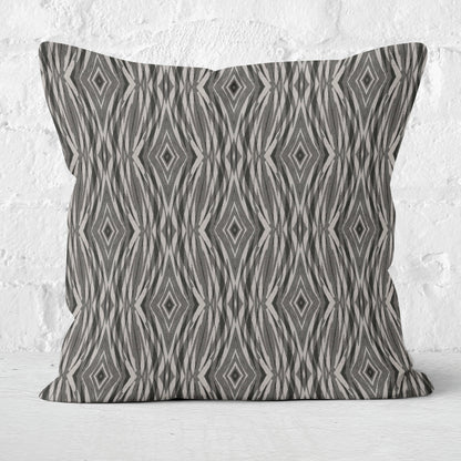 Square throw pillow featuring an abstract pattern in grey and neutral tones leaning against a white brick wall