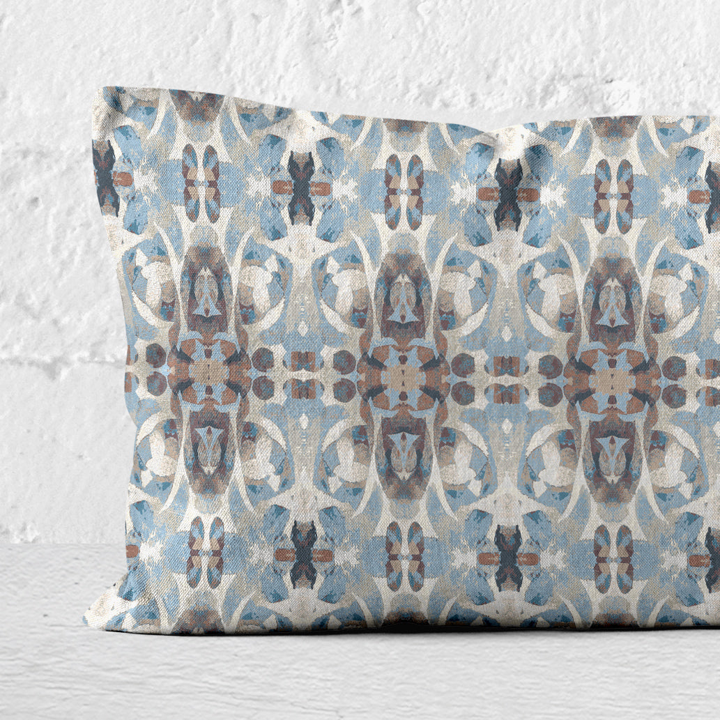 Detail of a 12 x 24 lumbar pillow featuring an abstract hand-painted blue and brown pattern leaning against a white brick wall.