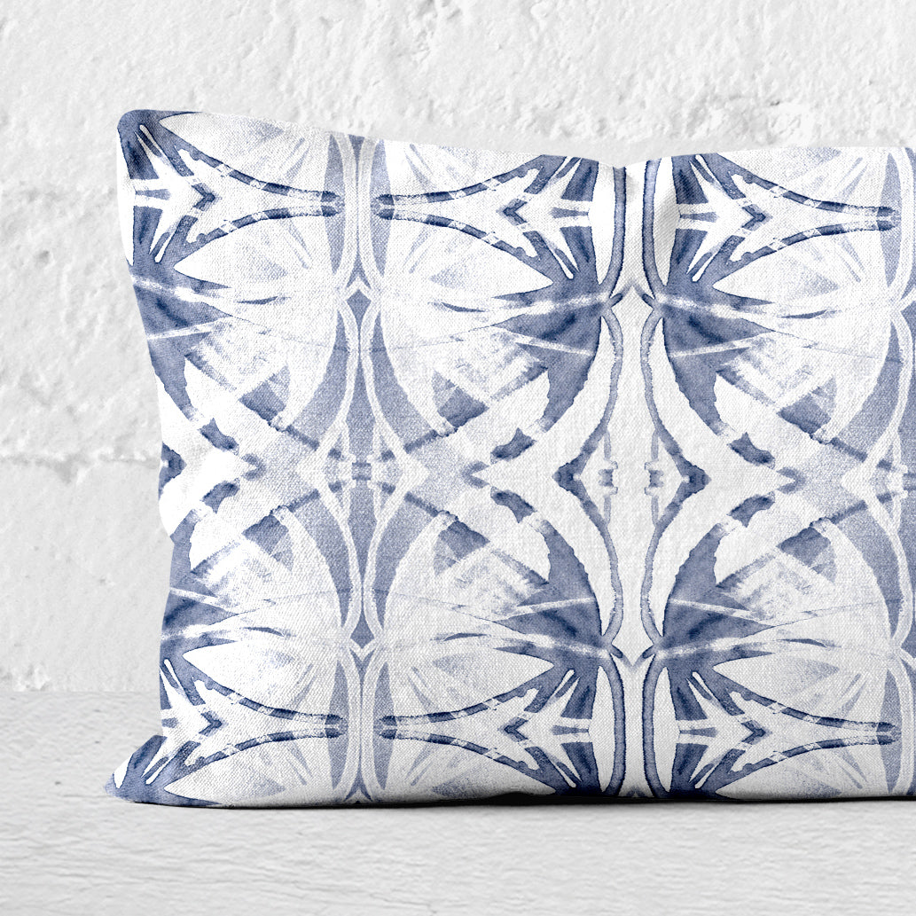 Detail of a rectangular pillow featuring abstract painted pattern in light blue and white set against a white brick wall.