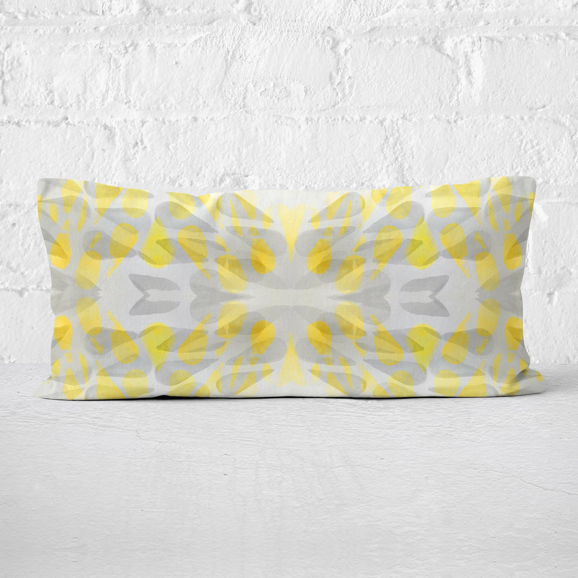 Rectangular lumbar pillow featuring an abstract hand-painted pattern in yellow and grey tones leaning against a white brick wall.