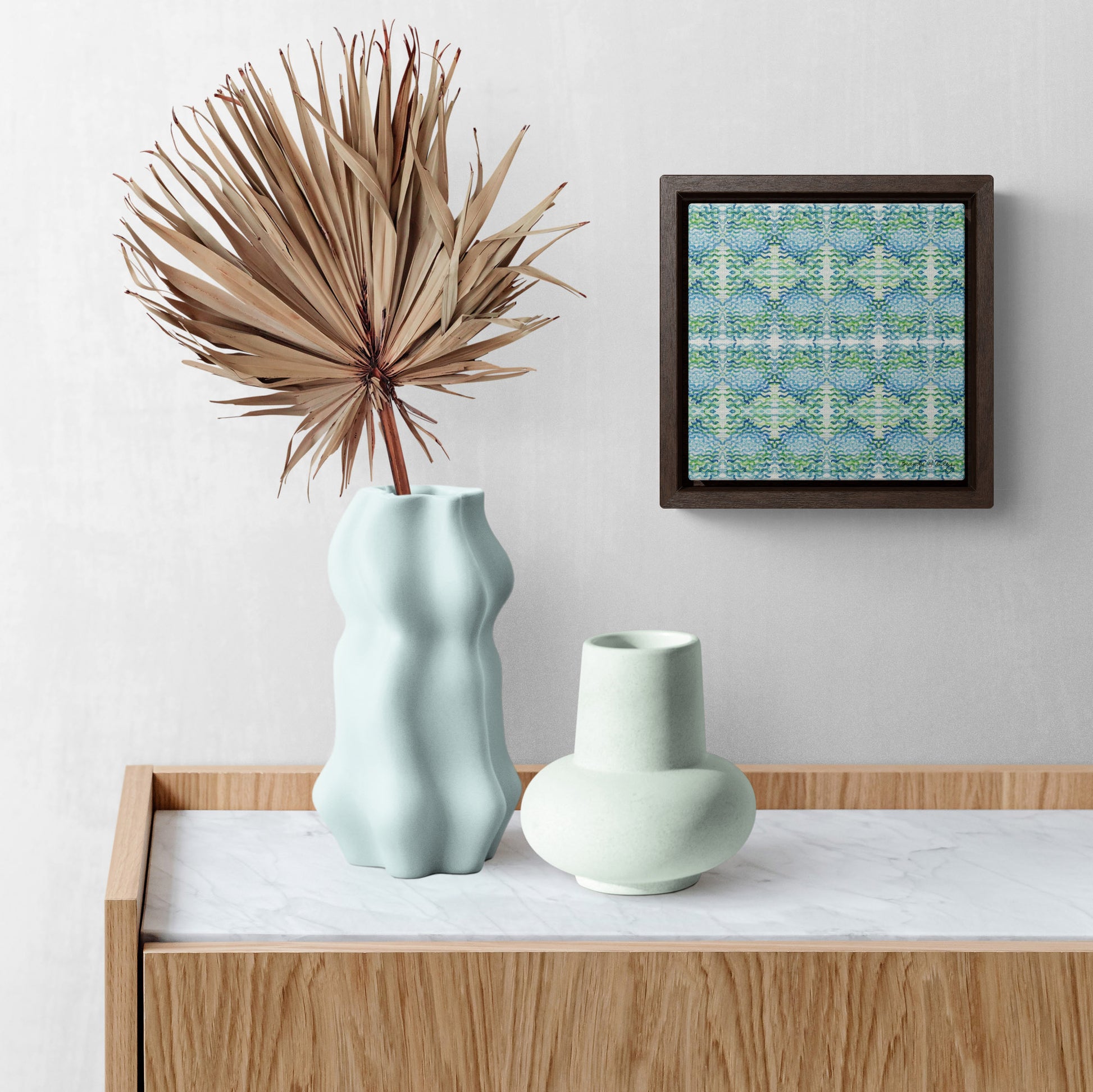 Stretched canvas featuring a blue and green abstract pattern in a black float frame, hanging on above blue vases with a palm frond