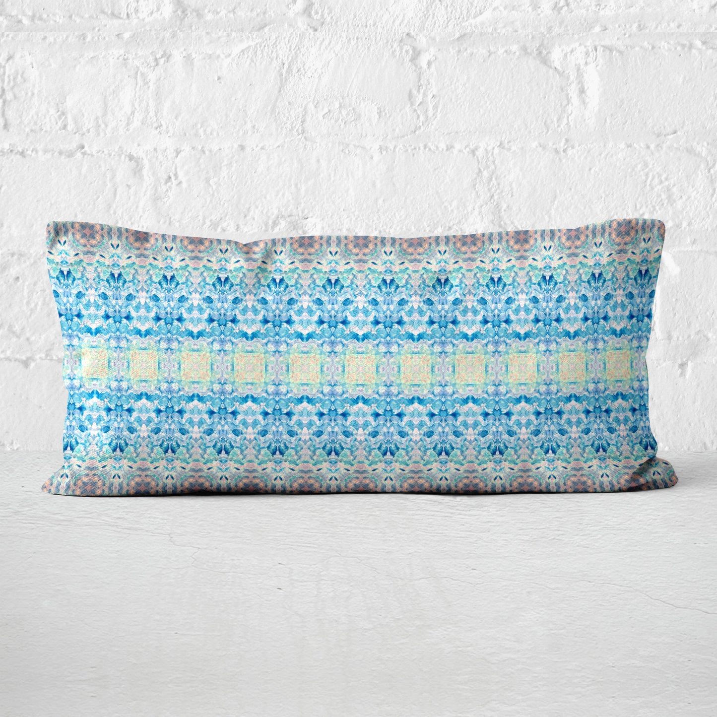Rectangular lumbar pillow featuring a handpainted pattern in blue, pink, and yellow leaning against a white brick wall.