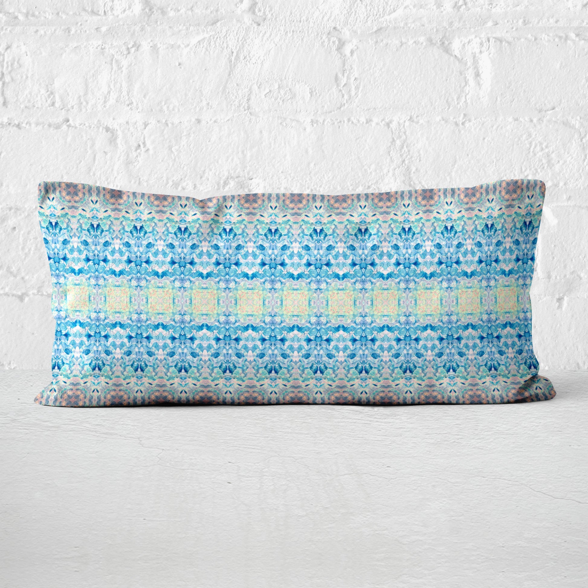 Rectangular lumbar pillow featuring a handpainted pattern in blue, pink, and yellow leaning against a white brick wall.