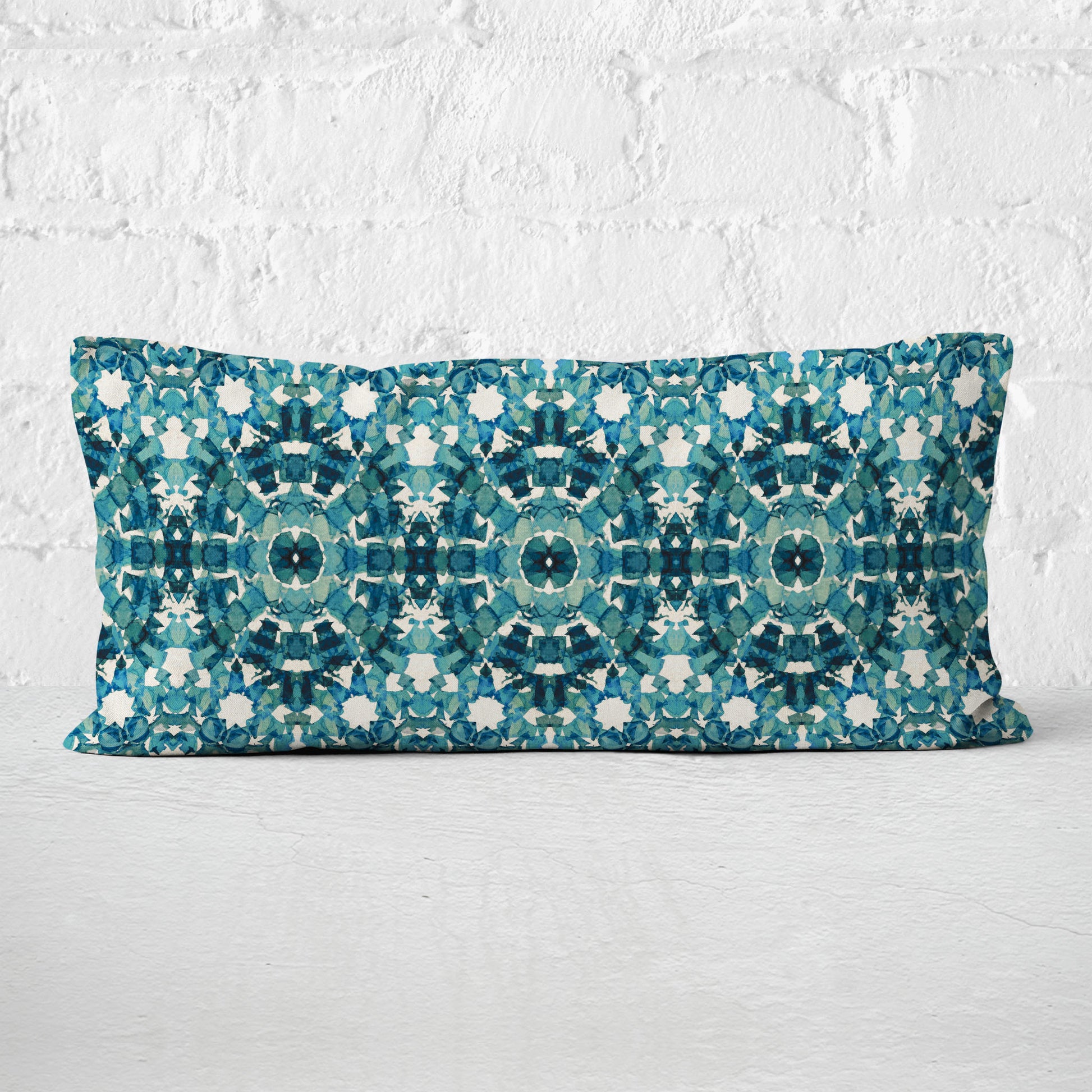 Rectangular lumbar pillow featuring handprinted pattern in teal leaning against a white brick wall.