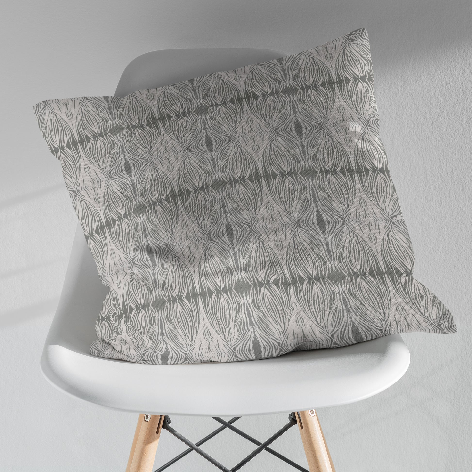 Square throw pillow featuring an abstract linocut pattern in grey and sitting on a white modern chair
