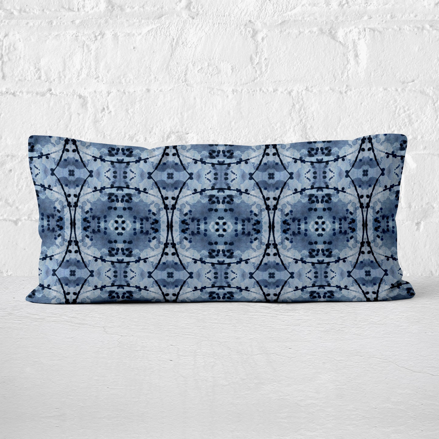 Rectangular lumbar pillow featuring an abstract hand-painted pattern in blue and black India Ink leaning against white brick wall