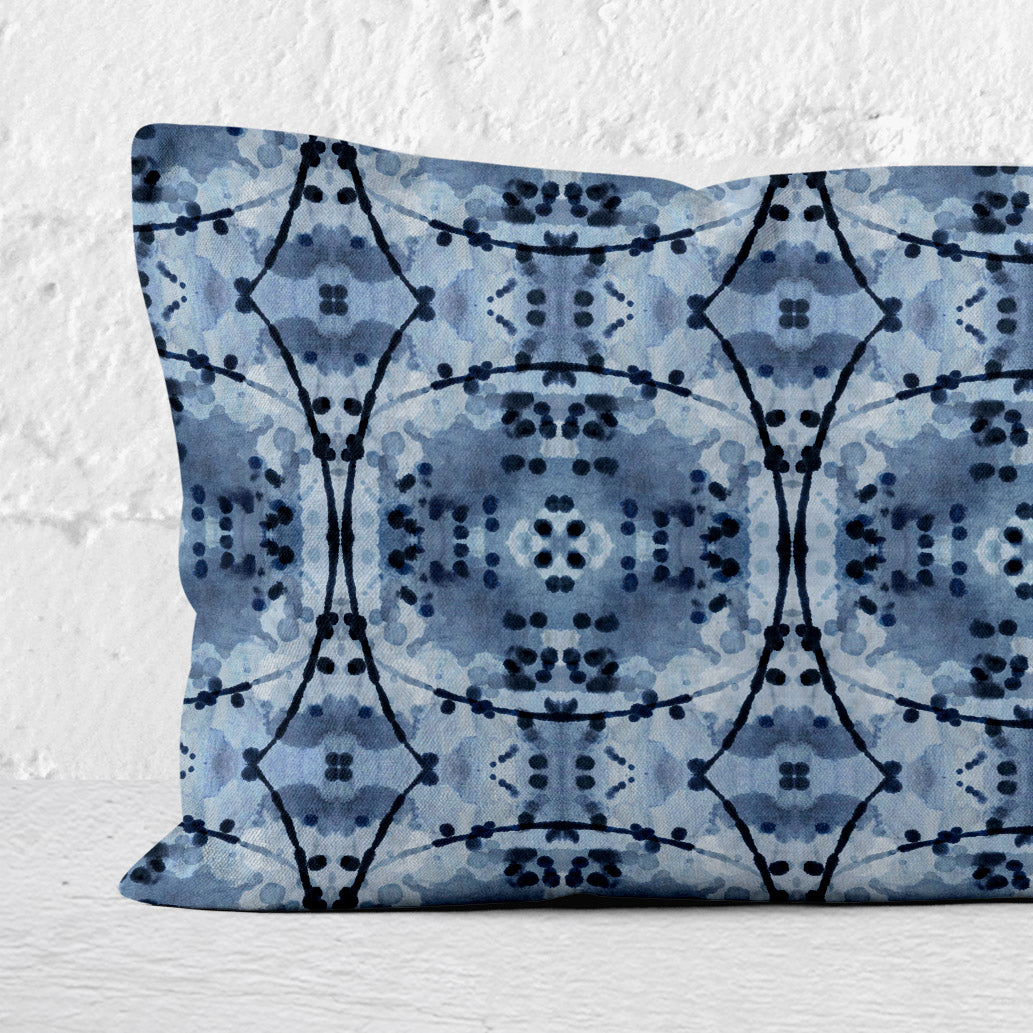 Detail of rectangular lumbar pillow featuring an abstract hand-painted pattern in blue and black India Ink leaning against white brick wall
