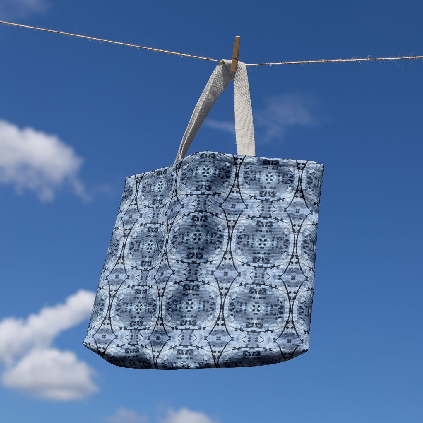 Large tote bag featuring an abstract hand-painted blue design, haniging on a clothesline in front of blue eky