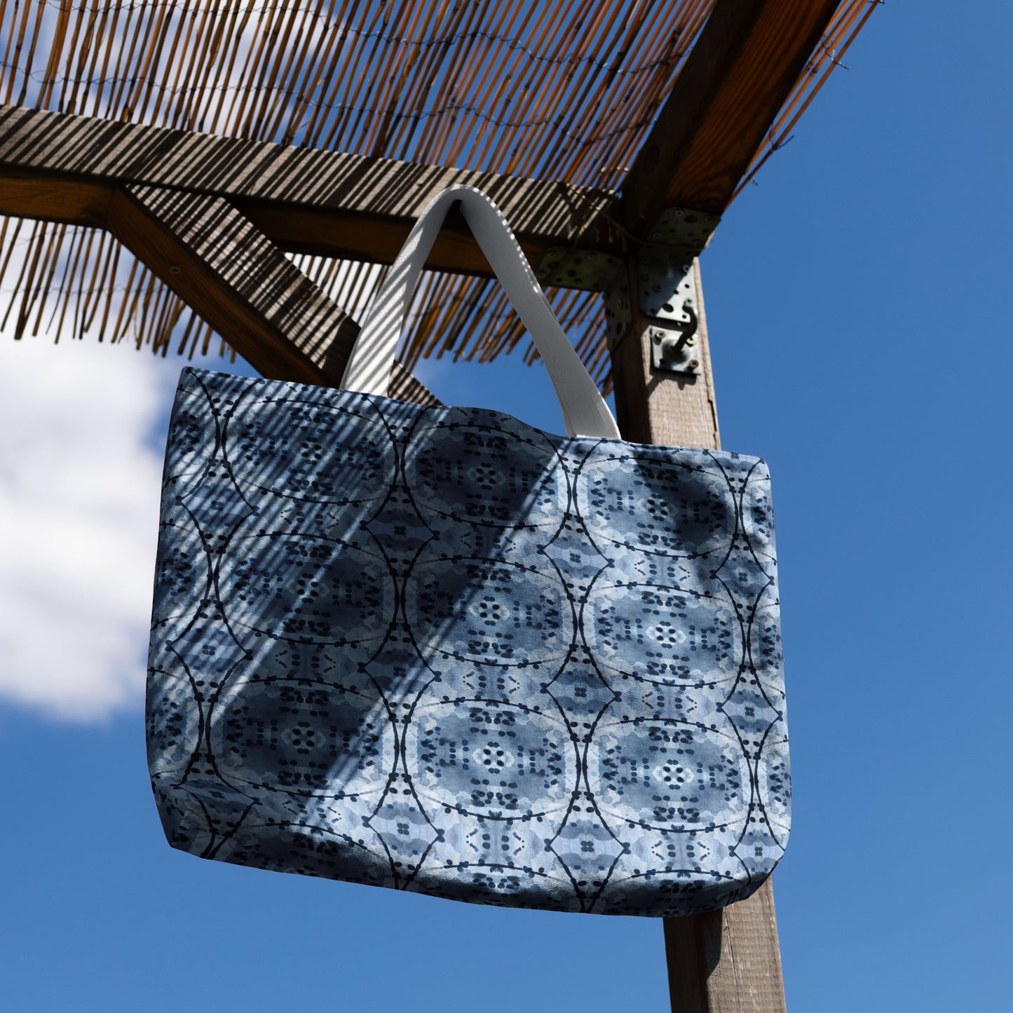 Large tote bag featuring an abstract hand-painted blue design, haniging from a potion cover  in front of blue eky