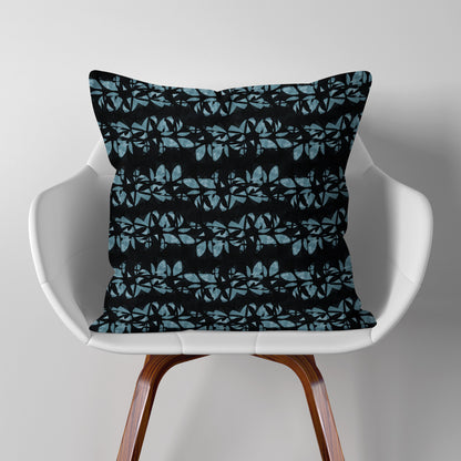 Square throw pillow featuring hand painted, abstract floral lei pattern in aqua blue and black, sitting on a modern white chair