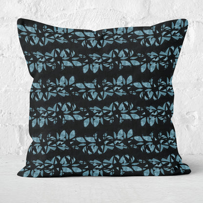 Square throw pillow featuring hand painted, abstract floral lei pattern in aqua blue and black, with a white brick wall in the background