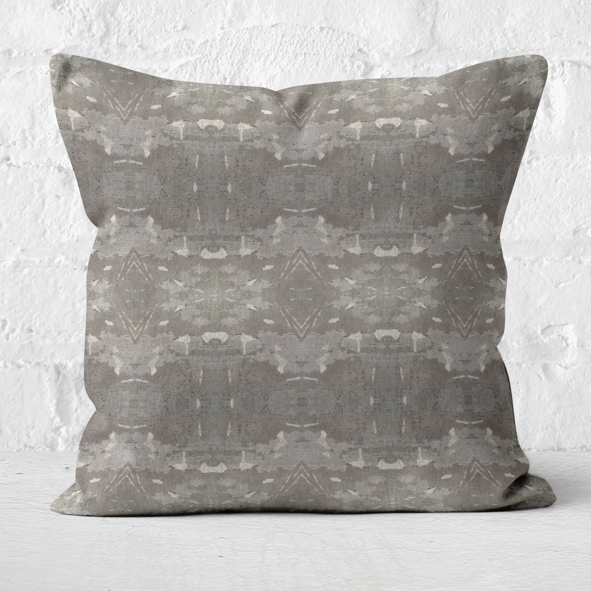 Square throw pillow featuring an abstract gray and khaki pattern, sitting in from of a white brick wall