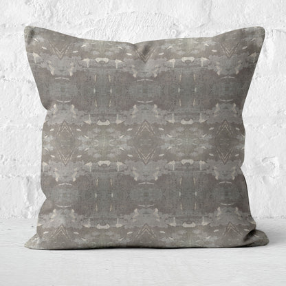 Square throw pillow featuring an abstract gray and khaki pattern, sitting in from of a white brick wall