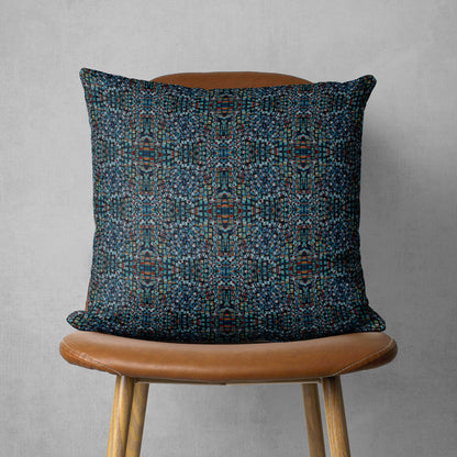 Square throw pillow featuring abstract hand-painted pattern in black, dark blue, and brown tones sitting on a brown chair