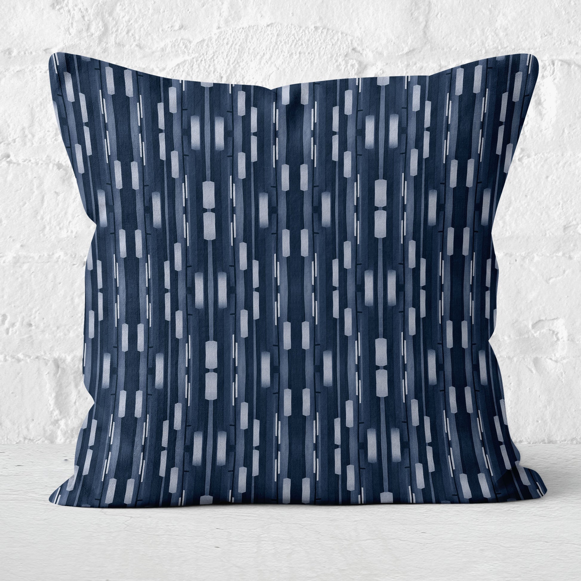 Pillow featuring collaged stripe pattern in blue and white with a white brick wall in background