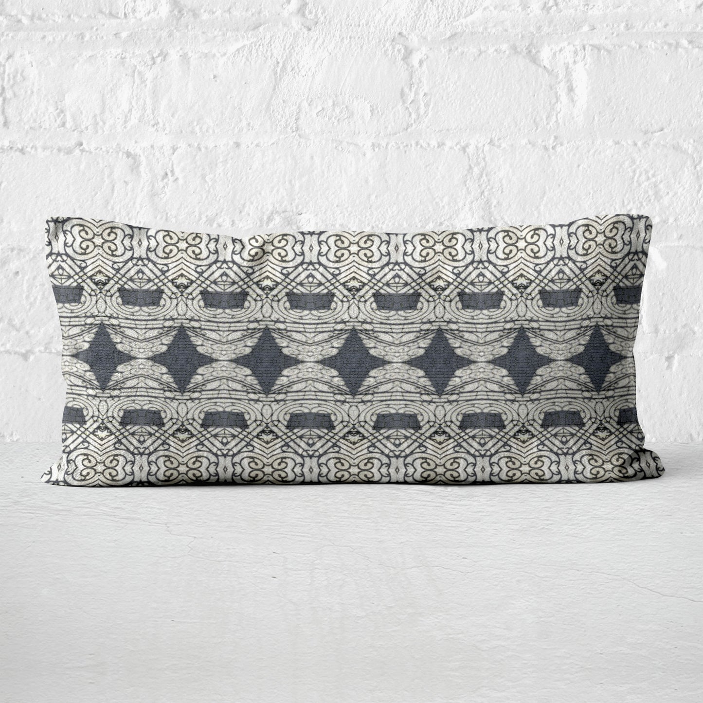 Rectangular lumbar pillow featuring abstract ornate pattern in black and grey tones leaning against white brick wall.