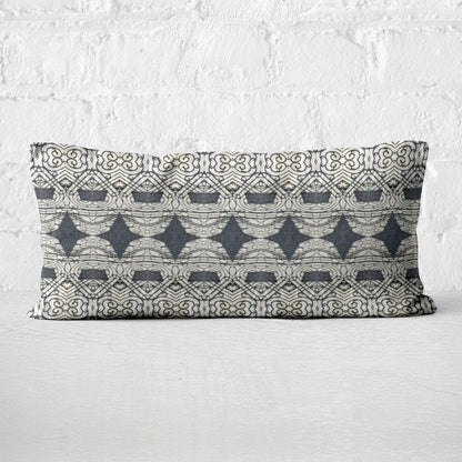 Rectangular lumbar pillow featuring abstract ornate pattern in black and grey tones leaning against white brick wall.