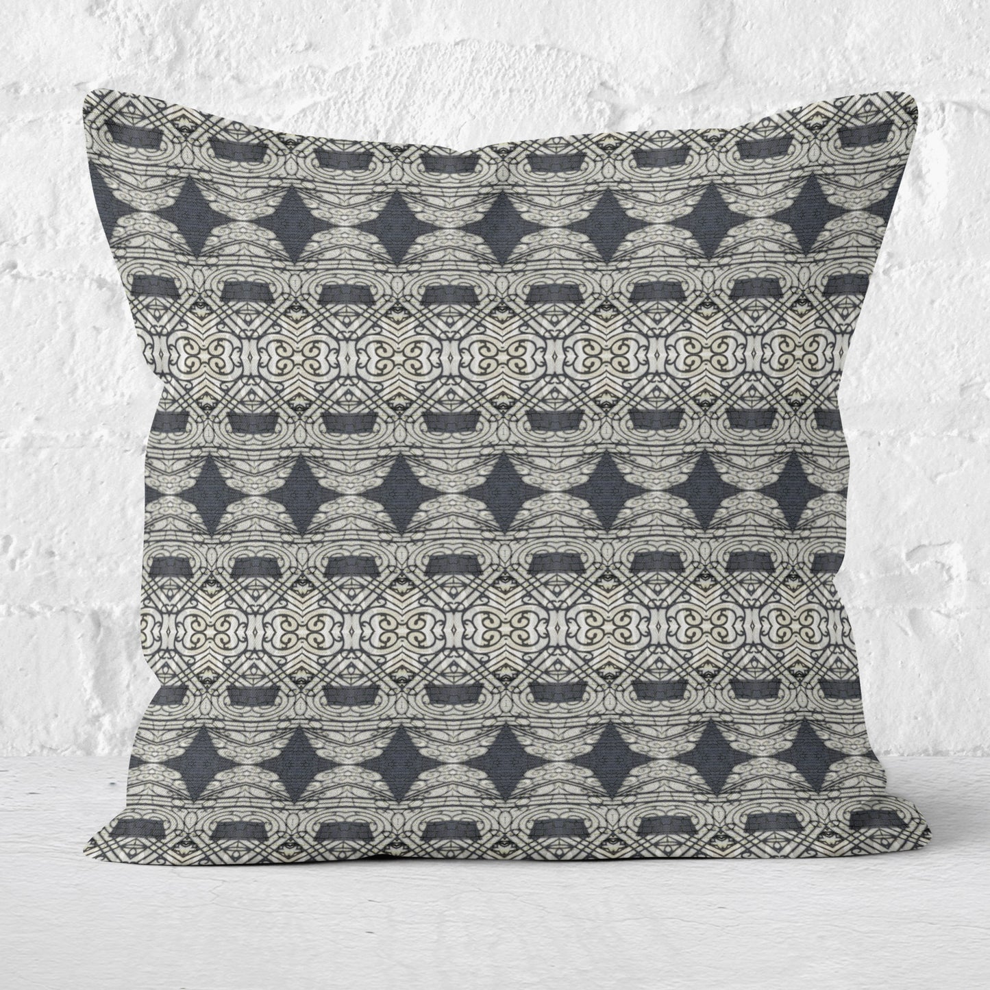 Square throw pillow featuring an abstract and ornate pattern in black and grey tones sitting in front of a white brick wall