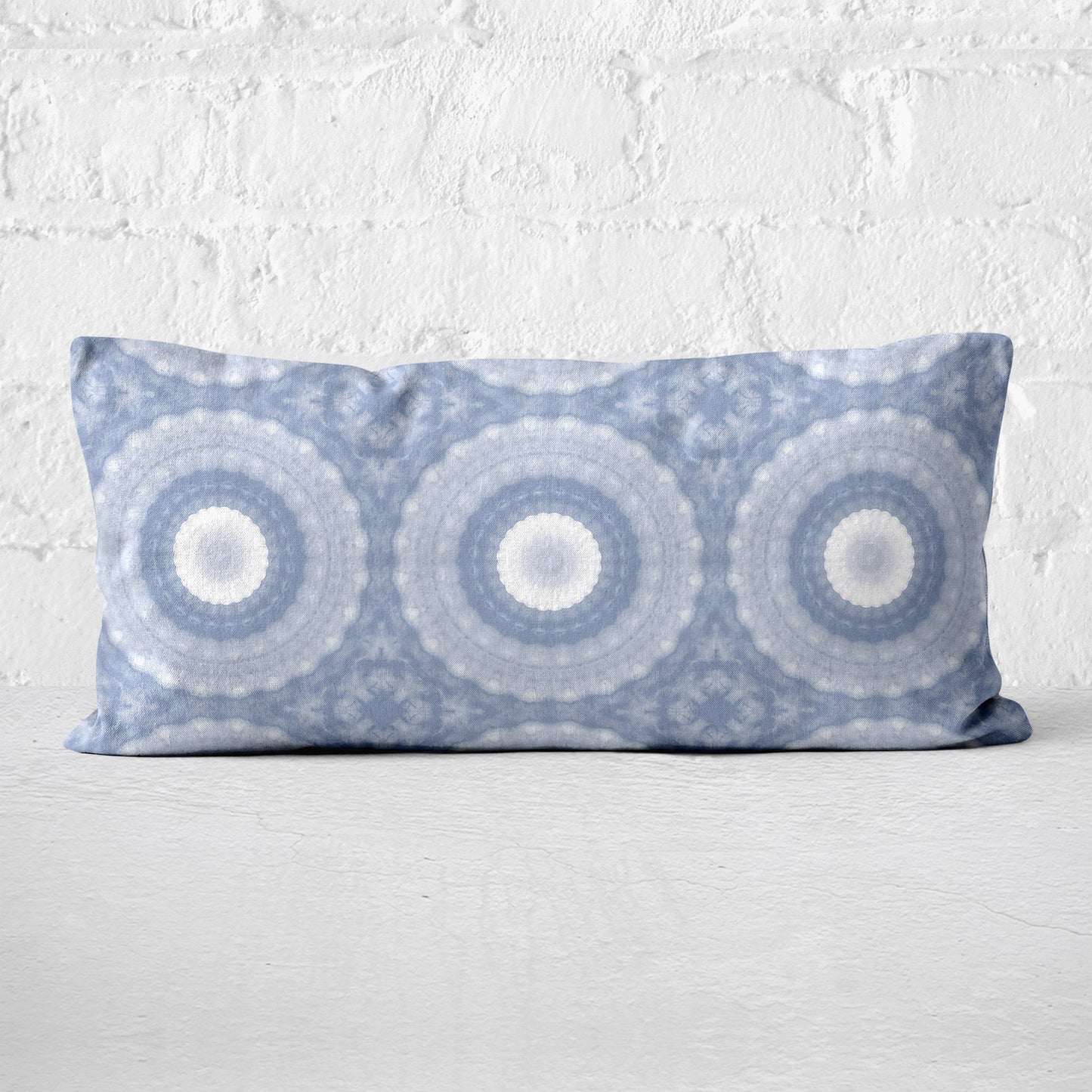 Lumbar pillow featuring a hand-painted circular pattern in periwinkle blue leaning against a white brick wall.