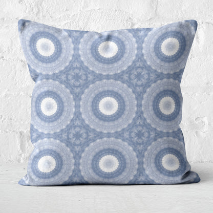 Square throw pillow featuring a periwinkle blue, purple, and white abstract circular pattern, standing in front of a white brick wall
