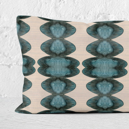 Detail of a rectangular lumbar pillow featuring abstract hand-painted pattern in blue, black, and cream leaning against a white brick wall.