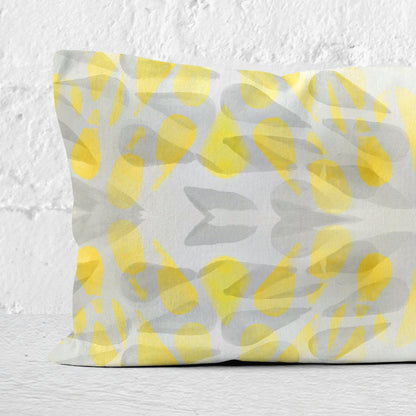Detail of a rectangular lumbar pillow featuring an abstract hand-painted pattern in yellow and grey tones leaning against a white brick wall.