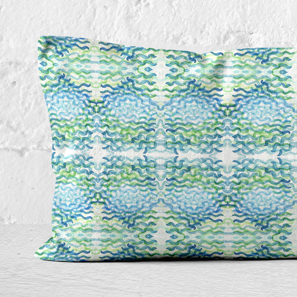 Detail of 12x24 lumbar pillow featuring an abstract hand-painted pattern in green and blue leaning against a white brick wall.