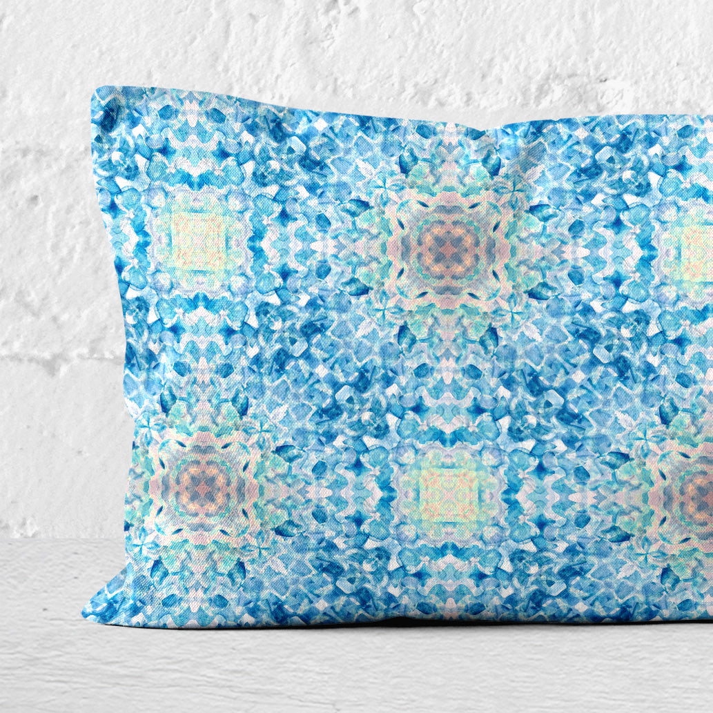 Detail of Rectangular lumbar pillow featuring a handpainted pattern in blue, pink, and yellow against a white brick wall.