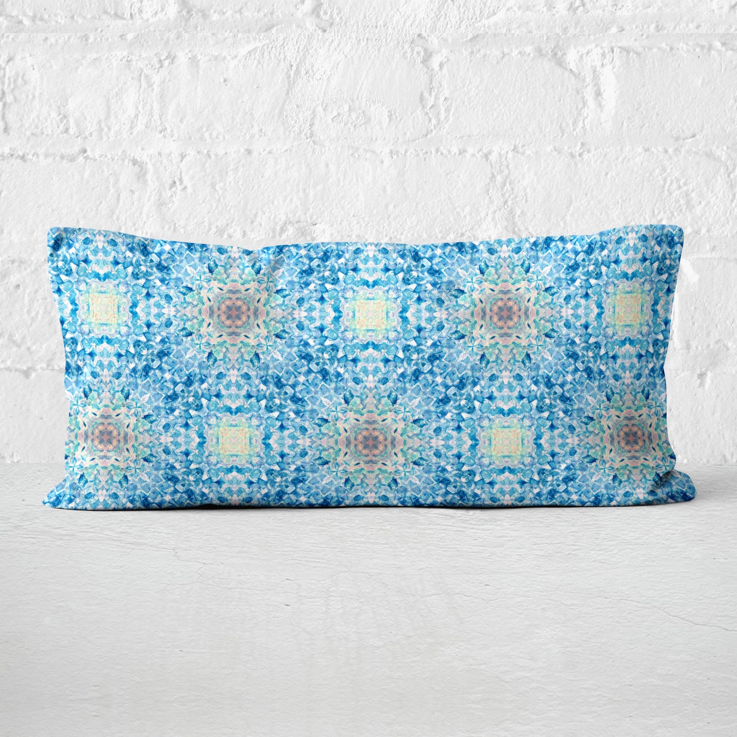 Rectangular lumbar pillow featuring a handpainted pattern in blue, pink, and yellow against a white brick wall.