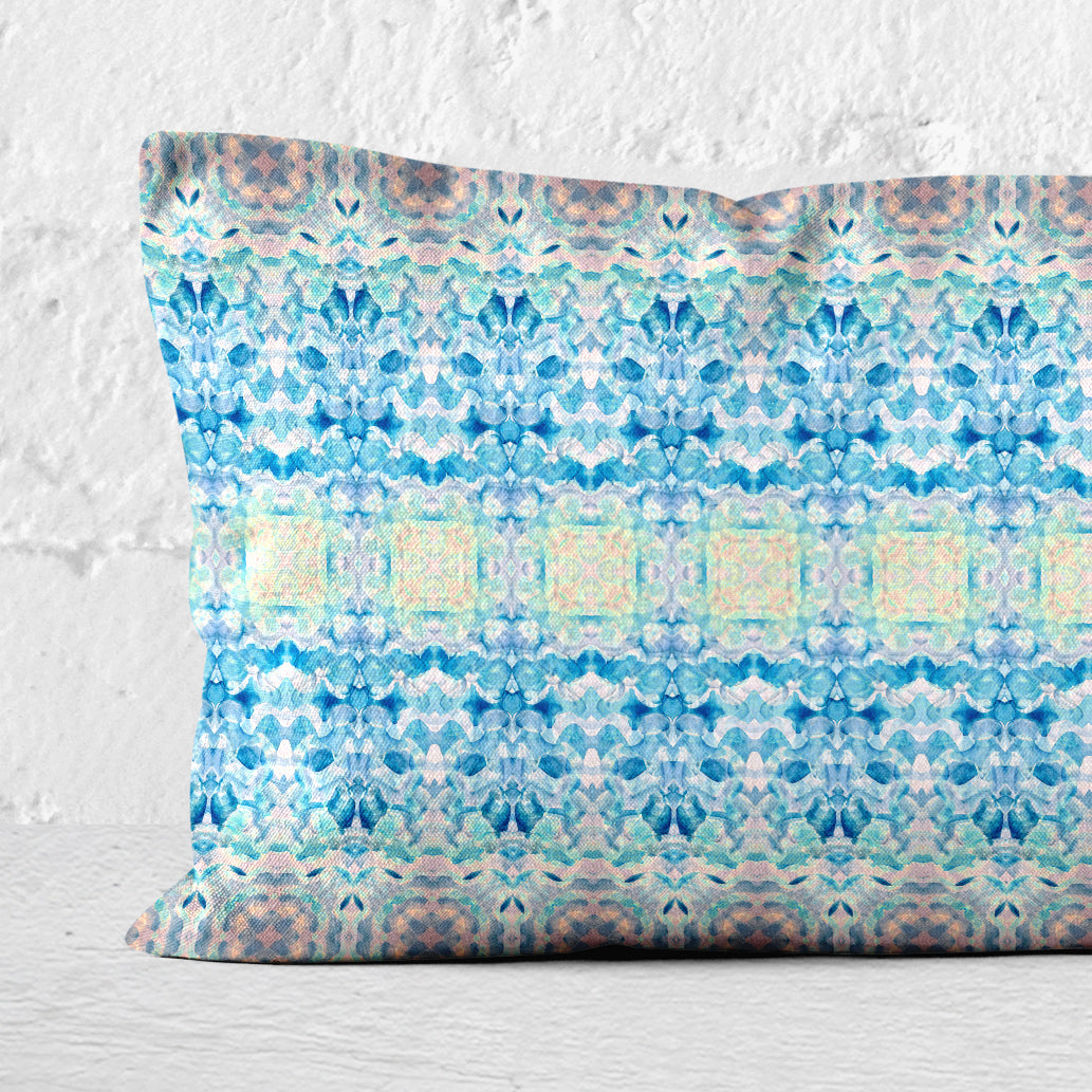 Detail of a rectangular lumbar pillow featuring a handpainted pattern in blue, pink, and yellow leaning against a white brick wall.