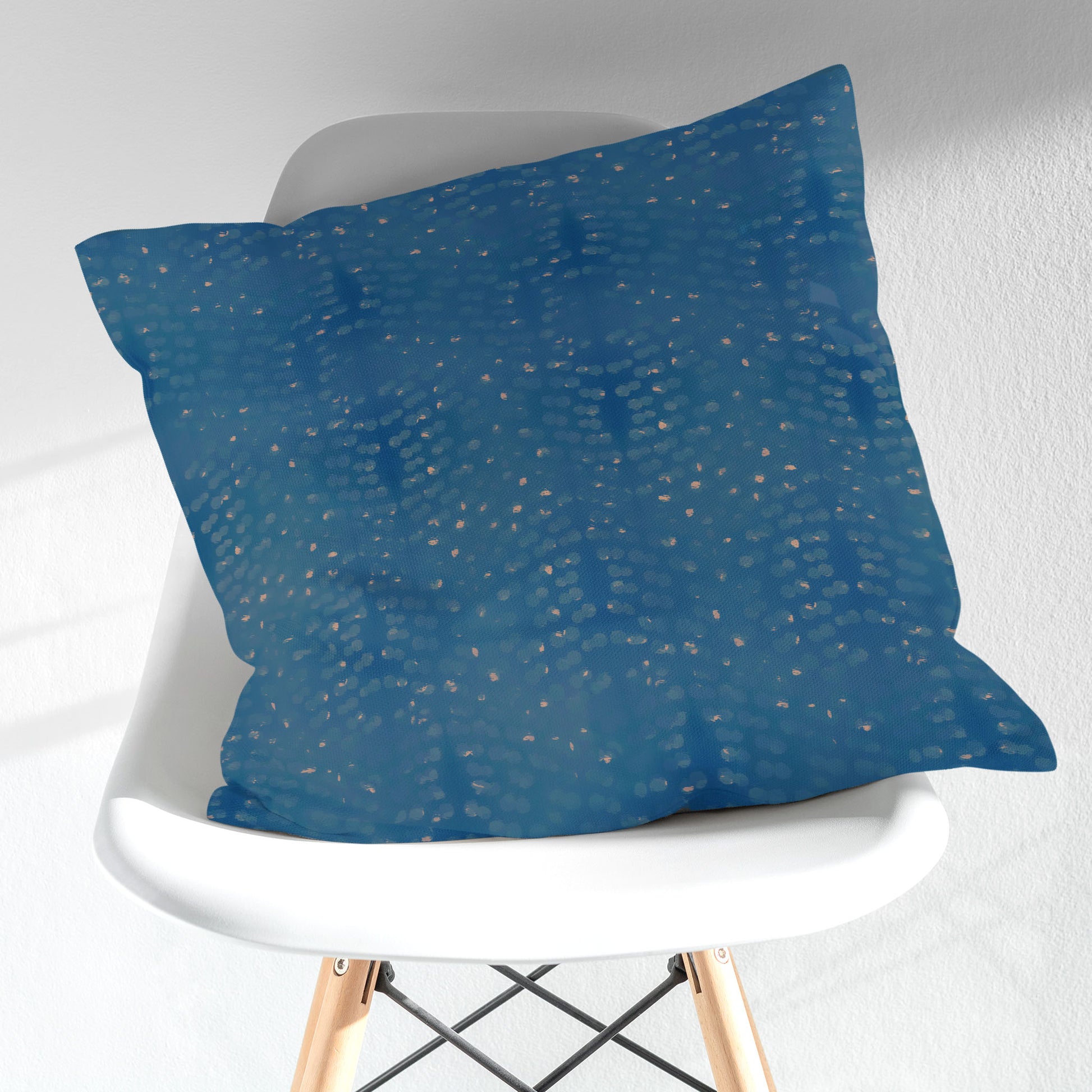 Square throw pillow featuring abstract hand painted pattern in blue and tan sitting on a modern style chair.