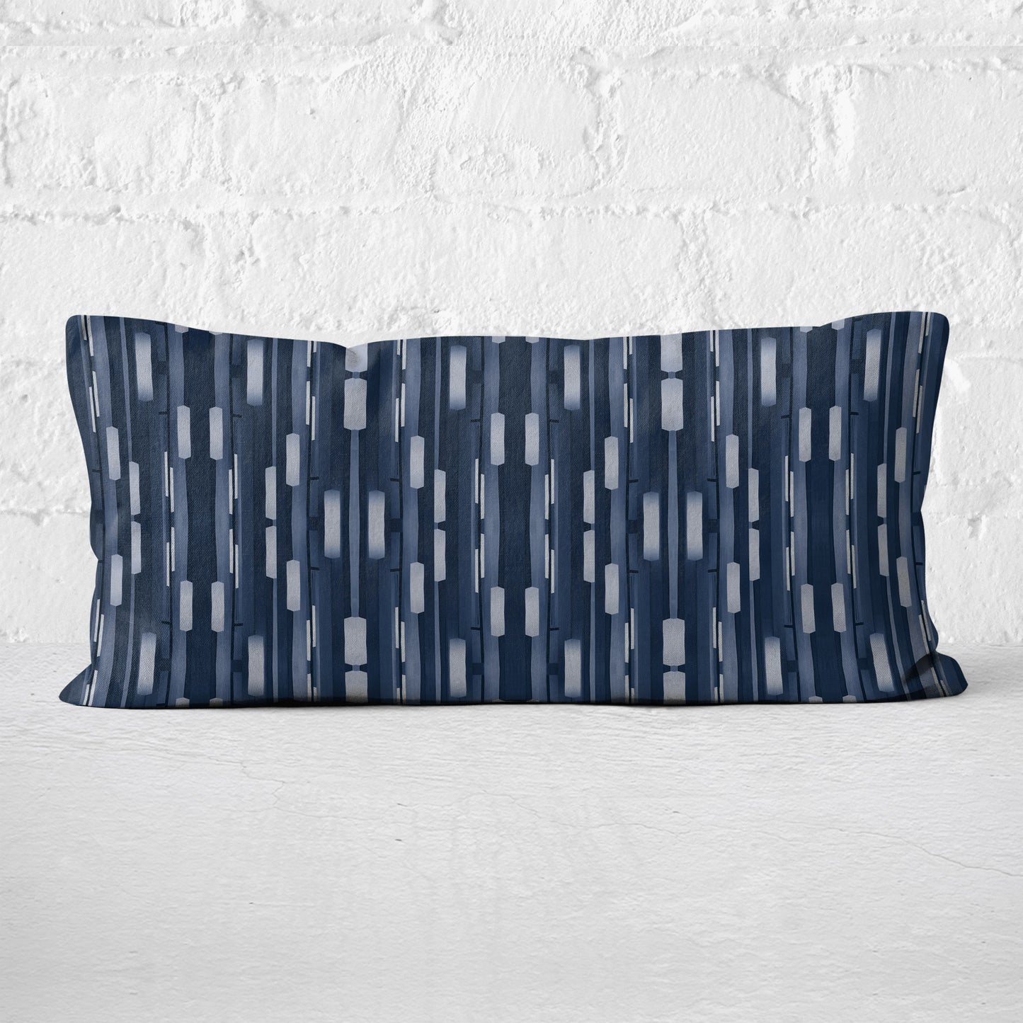 Rectangular lumbar pillow featuring a collaged stripe pattern in blue and white leaning against a white brick wall.