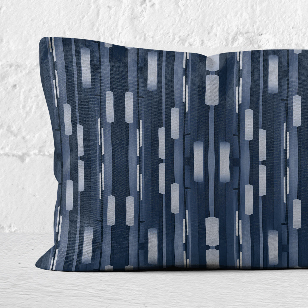 Detail of a rectangular lumbar pillow featuring a collaged stripe pattern in blue and white leaning against a white brick wall.