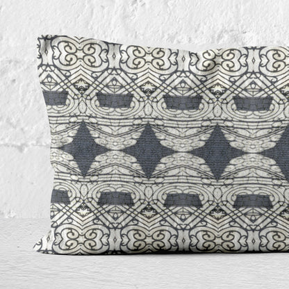 Detail of rectangular lumbar pillow featuring abstract ornate pattern in black and grey tones leaning against white brick wall.