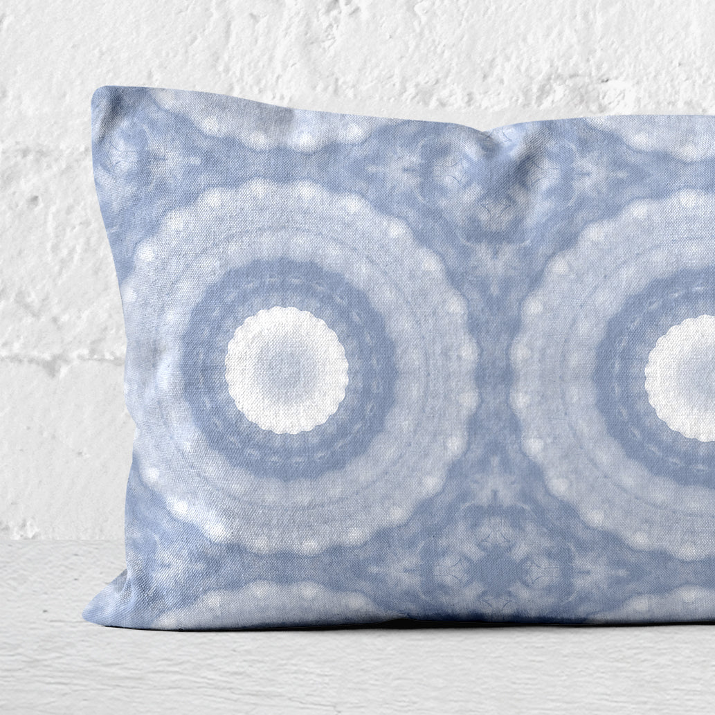 Detail of a 12x24 lumbar pillow featuring a hand-painted circular pattern in periwinkle blue leaning against a white brick wall.